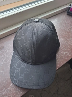 Svart keps från Gucci med mesh - Snygg svart keps från Gucci med klassiskt GG-mönster över hela kepsen. Framsidan är i tyg och baksidan har mesh för extra ventilation. Perfekt accessoar för en trendig streetstyle-look.