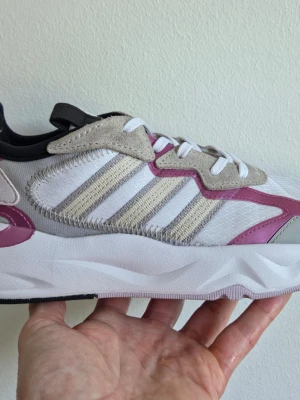 Adidas sneakers vit/grå/rosa - Snygga Adidas sneakers i vitt med grå och rosa detaljer. Skorna har klassiska tre ränder på sidan, mockaliknande partier och meshmaterial för extra komfort. Tjock vit sula och vita skosnören. Perfekta för en trendig streetstyle-look.