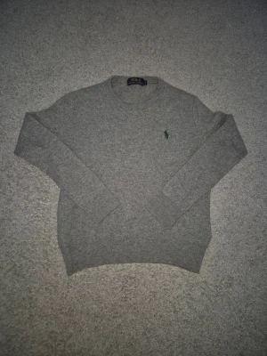 Polo Ralph Lauren - Klassisk grå stickad tröja från Polo Ralph Lauren med rund halsringning och den ikoniska gröna broderade loggan på bröstet. Tröjan har långa ärmar och ribbade muddar vid ärmslut och nederkant. Perfekt för en stilren och avslappnad look.