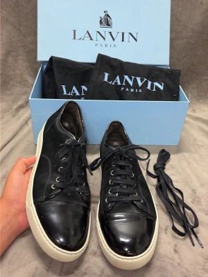 Svarta Lanvin sneakers i mocka och läder - Stilrena svarta sneakers från Lanvin med ovandel i mocka och glansiga tåpartier i läder. Skorna har vita sulor, svarta skosnören och klassisk låg profil. Perfekta för dig som gillar exklusiv och tidlös design.