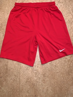 Röda sportshorts från Nike - Röda sportiga shorts från Nike med elastisk midja och vit broderad logga på vänster ben. Tillverkade i ett lätt och snabbtorkande material som passar perfekt för träning eller chill. Klassisk rak passform och enkel design. Passar XL för dam och M för herrar 