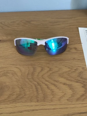 Sportiga vita solglasögon - Säljer ett par sportiga solglasögon med vit båge och spegelblå linser. De har en svepande, aerodynamisk form som passar perfekt för utomhusaktiviteter. Snyggt futuristisk look och riktigt bekväma att bära.