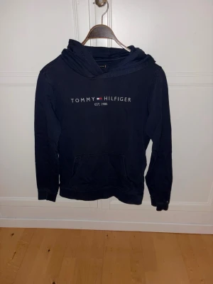 Mörkblå hoodie Tommy Hilfiger - Snygg mörkblå hoodie från Tommy Hilfiger med broderad logga och text 'EST. 1985' på bröstet. Klassisk känguruficka framtill och huva. Perfekt för en avslappnad och stilren look.
