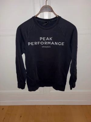 Mörkblå sweatshirt Peak Performance - Klassisk mörkblå sweatshirt från Peak Performance med vit logga och text på bröstet. Tröjan har rund halsringning, långa ärmar och ribbade muddar vid ärmslut och nederkant. Perfekt för en avslappnad och sportig stil. 