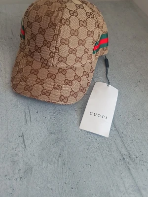 Beige keps från Gucci med GG-mönster - Snygg beige keps från Gucci med klassiskt GG-monogram över hela kepsen. På sidorna finns gröna och röda ränder som ger en ikonisk touch. Justerbar rem bak i läder och insida med svart tyg. Perfekt för dig som gillar exklusiva accessoarer.