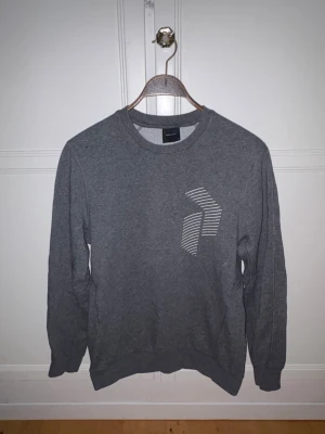 Grå sweatshirt Peak Performance - En stilren grå sweatshirt med rund halsringning och ett vitt grafiskt tryck på bröstet. Tröjan har långa ärmar och ribbade muddar vid ärmslut och nederkant. Perfekt för en avslappnad look. 