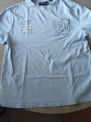 Ljusblå Supply and Demand t-shirt med tryck - Ljusblå t-shirt med coola tryck på både fram- och baksidan. Framsidan har vita och svarta detaljer med text och stjärnor, medan baksidan har ett stort 'supply'-tryck i gotisk stil. Tillverkad i mjuk bomull och har en avslappnad passform.