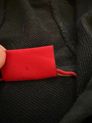 Svart hoodie från HUGO med röd logga - Säljer en svart hoodie från HUGO med stor röd logga på bröstet. Hoodien har huva med dragsko, magficka och ribbade muddar. Tillverkad i mjukt bomullstyg som är skönt att ha på sig. Perfekt för dig som gillar stilrena och klassiska plagg.