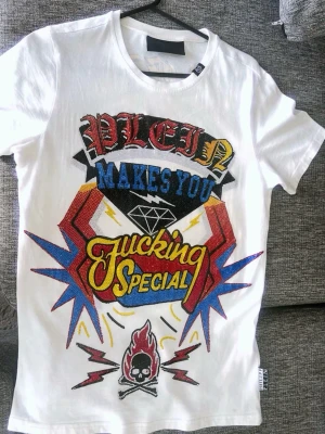 Vit t-shirt med färgglad paljettdekor - Säljer en vit t-shirt (märke) philipp plein med stort, färgglatt tryck och paljetter framtill. Texten 'PLEIN MAKES YOU FUCKING SPECIAL' och coola detaljer som blixtar, dödskalle och eld. Kortärmad modell i bomull med slim passform. Perfekt för dig som vill sticka ut.