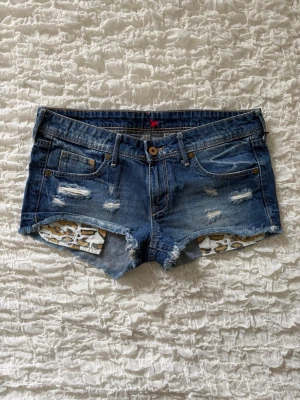 Blå jeansshorts med leoparddetaljer - Snygga blå jeansshorts med slitna detaljer och råa kanter. Insidan av fickorna har ett coolt leopardmönster som syns framifrån. Klassisk femficksmodell med knapp och dragkedja. Perfekta för sommaren och ger en edgy vibe till din outfit. Storlek 38 (alla mått finns i bilderna) 💕