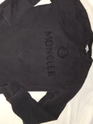 Moncler Sweatshirt, bra skick, M - Cool svart sweatshirt från Moncler med broderad logga och text framtill. Tröjan har rund halsringning, långa ärmar och ribbade muddar vid ärmslut och nederkant. Perfekt för en avslappnad och stilren look.