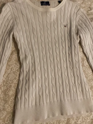 Vit tröja från Gant - Vit kabelstickad tröja från Gant med rund halsringning och broderad logga på bröstet. Tröjan har ribbade muddar vid ärmslut och nederkant, och är tillverkad i ett mjukt stickat material. Perfekt för en klassisk och stilren look.