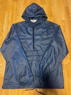 Mörkblå vindjacka från Lacoste - Snygg mörkblå vindjacka från Lacoste med huva och halv dragkedja framtill. Jackan har det klassiska krokodilmärket på bröstet, elastiska muddar och dragsko nedtill. Tillverkad i ett lätt och glansigt material som står emot vind.