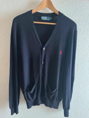  Ralph Lauren cardigan  - Klassisk mörkblå cardigan med V-ringning och knäppning framtill från Polo Ralph Lauren. Tröjan har två fickor, ribbade muddar och den ikoniska röda loggan broderad på bröstet. Perfekt att slänga över en t-shirt för en clean look.