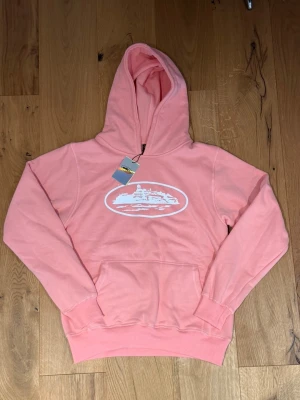 Rosa hoodie med vit tryck - Snygg rosa hoodie med vit tryck framtill som föreställer en byggnad. Hoodien har huva och en stor magficka. Muddar vid ärmslut och nederkant ger en skön passform. Perfekt för en avslappnad och trendig stil.