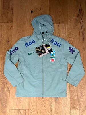 Mintgrön Brasilien vindjacka med huva  - Mintgrön vindjacka med huva och dragkedja framtill. Jackan har Brasilien CBF-logga, grönt Nike-märke och flera tryck i blått och lila på bröst och ärmar. Tillverkad i GORE-TEX-material, perfekt för sportiga dagar. Flera sponsormärken syns på framsidan.