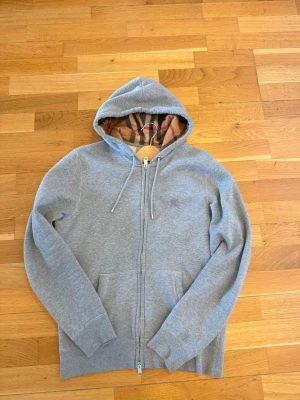 Grå zip hoodie från Burberry - Snygg grå hoodie från Burberry med dragkedja och klassiskt rutigt foder i huvan. Tröjan har fickor framtill och justerbar dragsko i huvan. Perfekt för dig som vill ha en stilren och exklusiv look med en touch av Burberrys ikoniska mönster.