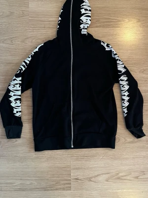 Svart hoodie med vit tribal-detalj - Storlek säger XXL men den passar som en S/M.  Svart hoodie med dragkedja och huva. Ärmarna och huvan har vita tribal-inspirerade mönster längs sidorna. Tillverkad i mjukt material och har ribbade muddar vid ärmslut och nederkant. Perfekt för en streetwear-look.