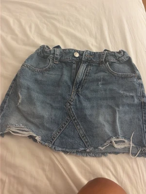 Blå jeanskjol med fransig kant - Säljer en blå jeanskjol i kort modell med fransig, rå kant nertill och coola detaljer framtill. Kjolen har fickor både fram och bak samt bälteshällor. Perfekt för dig som gillar en avslappnad och trendig stil.