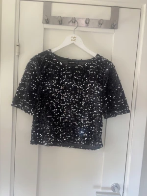 Svart paljettopp med kort ärm - Glittrig svart topp täckt med paljetter, perfekt för dig som vill sticka ut. Modellen är croppad med korta ärmar och rund halsringning. Snygg att styla till både jeans och kjol för en trendig look.