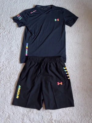 Svart träningsset från Under Armour - Snyggt svart träningsset från Under Armour med t-shirt och shorts. Setet har färgglada loggor och detaljer i regnbågsfärger på både tröja och byxa. Perfekt för gymmet eller löpning. Materialet är lätt och andas, vilket gör det bekvämt under träning.