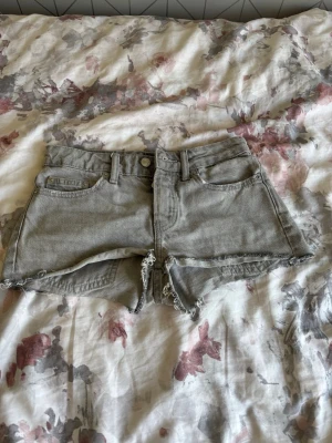 Grå lågmidjade denimshorts med fransig kant - Snygga grå denimshorts med rå, fransig kant och klassisk femficksdesign. Shortsen har bälteshällor och knappstängning framtill. Perfekta för varma dagar och en avslappnad stil.
