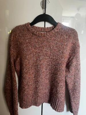 Rosa melerad stickad tröja Indiska - Mysig stickad tröja från Indiska i en rosa melerad färg med inslag av lila och orange. Tröjan har rund halsringning, långa ärmar och ribbade muddar vid ärmslut och nederkant. Perfekt för kyliga dagar och enkel att matcha med jeans eller kjol.