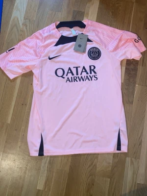 Rosa PSG matchtröja från Nike - Säljer en snygg rosa PSG matchtröja från Nike med svart logga och Qatar Airways-tryck på bröstet. Tröjan har korta ärmar, rund hals och detaljer i svart vid axlar och sidor. Perfekt för fotbollsträning eller att bära till vardags.