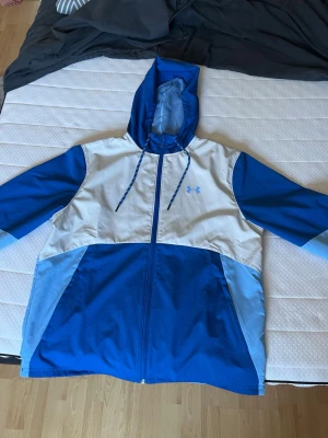 Blå och vit vindjacka från Under Armour - Snygg vindjacka från Under Armour i blått och vitt med dragkedja framtill och huva med dragsko. Jackan har flera blå nyanser och en diskret logga på bröstet. Perfekt för blåsiga dagar och sportiga outfits.