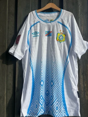 DR Kongo fotbollströja från Umbro - Säljer en vit fotbollströja från Umbro med DR Kongos landslagsemblem och flagga på bröstet. Tröjan har blå detaljer och grafiskt mönster nedtill, rund halsringning och korta ärmar. Perfekt för match eller träning!