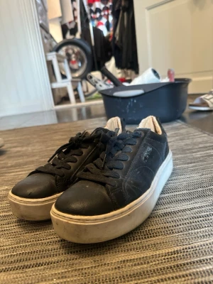 Svarta sneakers med vit sula - Säljer ett par svarta sneakers med tjock vit sula och svarta skosnören. Skorna har en klassisk låg modell och är tillverkade i ett slätt material som ser ut som läder. Perfekta för en clean och stilren look.