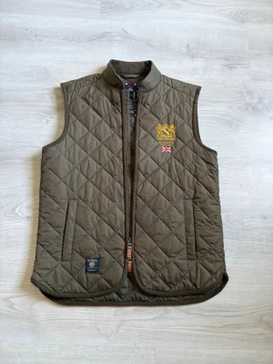 Olivgrön quiltad dunväst från Morris - Snygg olivgrön dunväst från Morris med quiltat mönster, ståkrage och dragkedja framtill. Västen har två fickor och detaljer som broderad logga och brittisk flagga på bröstet. Perfekt för lager på lager och stilrena outfits.