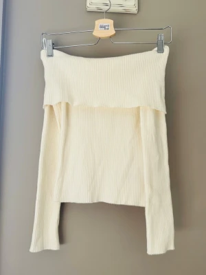 Off shoulder cremevit stickad tröja - Trendig off shoulder stickad tröja i cremevitt med ribbad struktur. Långa ärmar och bred, nedvikt kant upptill ger en snygg och avslappnad look. Perfekt att styla med jeans eller kjol för en modern vibe.