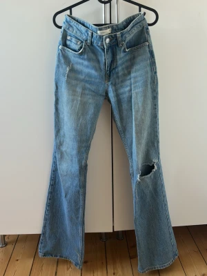 Blå bootcut jeans med hål - Säljer ett par ljusblå bootcut jeans med slitningar och ett stort hål på ena knät. Jeansen har en medelhög midja. Perfekta för en avslappnad och trendig look. 
