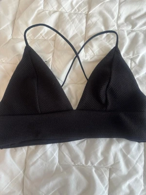 Svart bikiniöverdel med tunna band - Snygg svart bikiniöverdel med triangelformade kupor och tunna axelband som korsas i ryggen. Materialet har en lätt ribbad struktur som ger en modern känsla. Perfekt för stranden eller poolen.