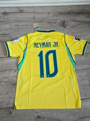 Brasilien Neymar Jr matchtröja 2026 - Säljer en gul Brasilien matchtröja från Nike med Neymar Jr och nummer 10 på ryggen. Tröjan har blå och gröna detaljer, korta ärmar och officiellt landslagsmärke på bröstet. Perfekt för dig som älskar fotboll och vill stötta Brasilien.