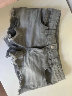 Gråa jeansshorts med fransig kant - Gråa jeansshorts med råa, fransiga kanter och coola detaljer som fickor med lock och knappar bak. Klassisk femficksmodell med knapp och dragkedja framtill. Perfekta för sommardagar och en avslappnad streetstil.