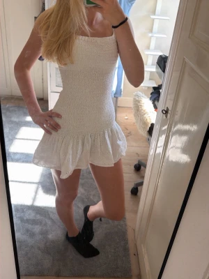Vit smockad sommarklänning - Supersöt vit klänning med smockad överdel och volangig kjol. Klänningen har tunna axelband och broderade blommor på kjolen. Perfekt för varma dagar och ger en fräsch och ungdomlig look.