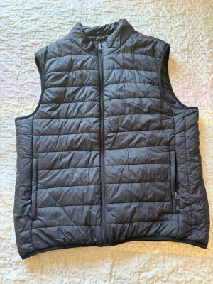 Svart quiltad dunväst - Snygg svart dunväst med quiltat mönster och dragkedja framtill. Västen har två sidofickor med dragkedja och hög krage. Perfekt att slänga över en hoodie eller tröja för extra värme. Materialet är lätt och smidigt, vilket gör den enkel att bära.