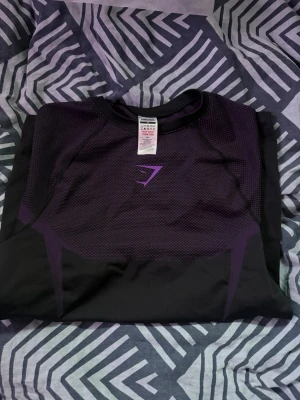 Gymshark Onyx V5 T-shirt Purple - Snygg svart och lila träningströja från Gymshark med meshdetaljer och logga på bröstet. Tröjan har rund halsringning och är tillverkad i ett stretchigt funktionsmaterial som andas, perfekt för gymmet eller löprundan.