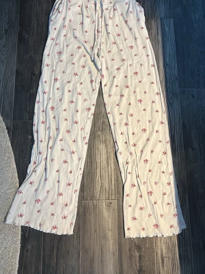 Vita pyjamasbyxor med blommor - Säljer ett par vita pyjamasbyxor med små rosa blommor och diskreta ränder. Byxorna har resår och snörning i midjan för skön passform. Perfekta för mysiga hemmakvällar eller lata morgnar.