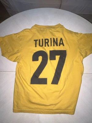 Gul AIK t-shirt Turina 27 - Säljer en gul AIK t-shirt med svart tryck, nummer 27 och namnet Turina på ryggen. Framsidan har AIK:s klubbmärke och ÅBRO-tryck. Perfekt för dig som vill hylla en AIK-legend. T-shirten har rund hals och korta ärmar.
