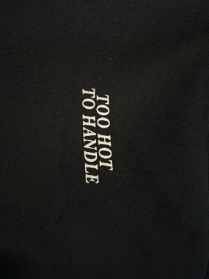 Svart hoodie med texttryck - Svart hoodie med vit text 'TOO HOT TO HANDLE' på bröstet. Klassisk känguruficka framtill och huva. Perfekt för dig som gillar streetwear och vill ha en enkel men snygg look. Mjuk och skön bomullskänsla.