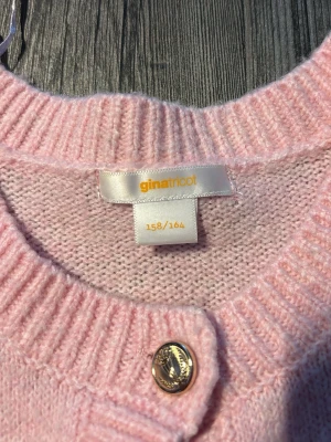 Ljusrosa kofta från Gina Tricot - Söt ljusrosa stickad kofta från Gina Tricot i storlek 158/164. Koftan har ribbade muddar och snygga guldfärgade knappar framtill. Perfekt för att addera en mjuk och trendig touch till din outfit.