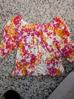 Blommig offshoulder blus i starka färger stl L - Säljer en superfin offshoulder blus med puffiga ärmar och smockdetaljer. Blusen har ett färgstarkt blommönster i rosa, orange och vitt. Materialet är tunt och luftigt, perfekt för sommaren. Passar dig som gillar färg och vill sticka ut!