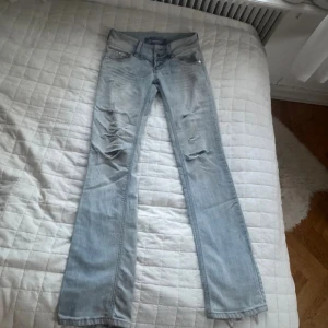 Ljusblå slitna bootcut jeans, RESERVERADE! - Säljer ett par ljusblå jeans med slitningar och hål på framsidan. Modellen är lite mer åt straight hållet och en av knapparna saknas men det syns inte så mkt när man knäpper den nedre knappen. Midjemåttet är 34cm och innerbenslängden är 77cm💕