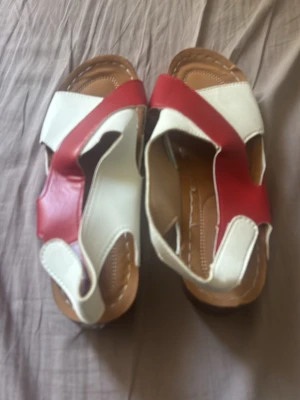 Röda och vita sandaler med breda remmar, storlek 41. - Säljer ett par sandaler med breda remmar i rött och vitt. De har öppen tå och häl, samt en bekväm brun sula. Perfekta för sommaren och ger en cool retrovibe till din outfit.