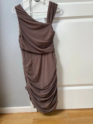 Brun asymmetrisk draperad klänning - Säljer en brun, asymmetrisk klänning med draperade detaljer och oneshoulder-design. Klänningen har en tight passform och är tillverkad i ett mjukt, stretchigt material som ger en snygg siluett. Perfekt för dig som vill sticka ut med en unik look. 