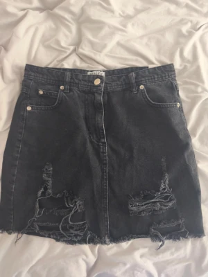 Svart jeanskjol från Madlady - Säljer en svart jeanskjol från Madlady i kort modell. Kjolen har slitna detaljer och fransig nederkant för en edgy look. Klassisk femficksdesign och knappstängning framtill. I storleken S