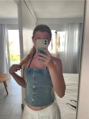 Ljusblå denim tubtopp med knappar - Trendig ljusblå tubtopp i denim med snygga silverfärgade knappar framtill och rå kant nedtill. Toppen är croppad och har en figurnära passform som ger en cool och avslappnad vibe. Perfekt för sommardagar och festivaler.